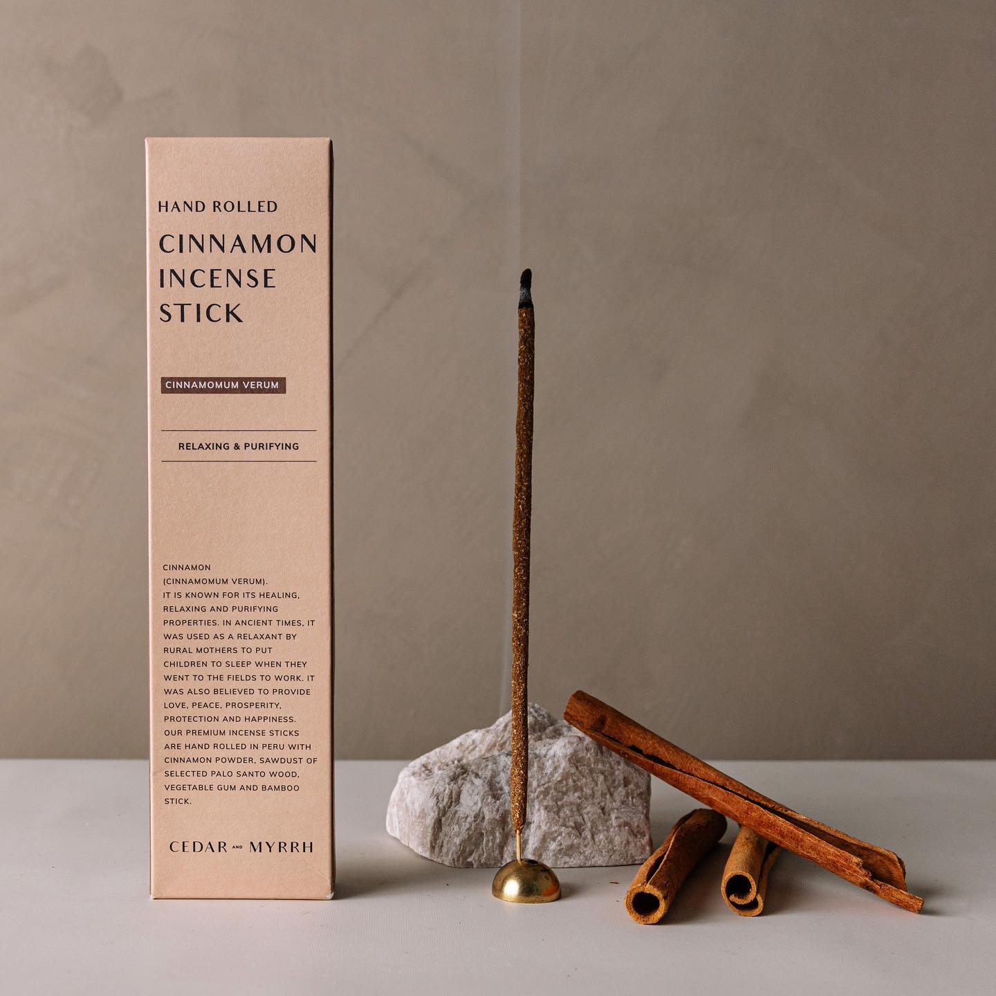 An incense stick in an incense holder that is lit and burning, along with its box and cinnamon sticks which the incense sticks are made with by Cedar and Myrrh.