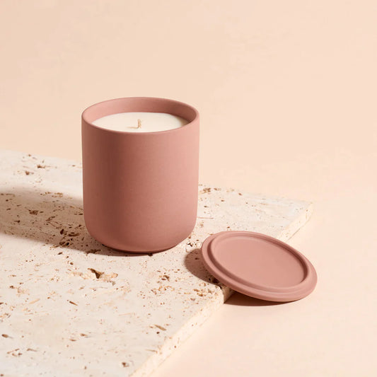 A candle in a rose pink ceramic vessel by Lit By drew, with the lid removed and placed on the side in damsum plum and patchouli scent.