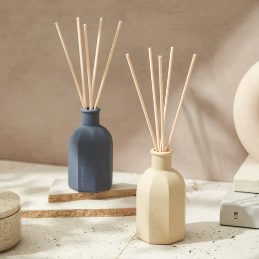 A reef diffuser in a navy blue and in a beige ceramic container with wooden reeds by Lit By Drew, sitting on a desk adding elegance to the decor and fragrance to the room.