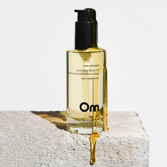 A bottle of body oil, bath oil sitting on a stone surface, with the oil dripping down the bottle. Made by Om Organics with coconut scent.