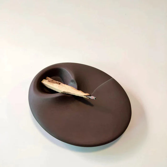 A palo santo holder in dark brown color, handmade with ceramic stoneware by Siesta Studio, Turkey, holding a palo santo stick that is burning.